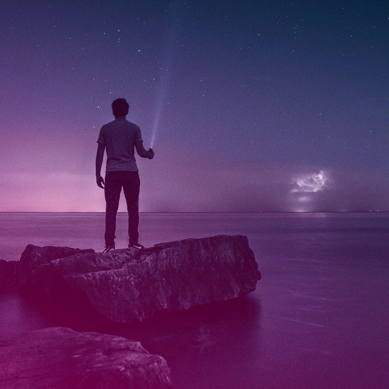 a man holding a flashlight standing on a rock overlooking the sea at dusk